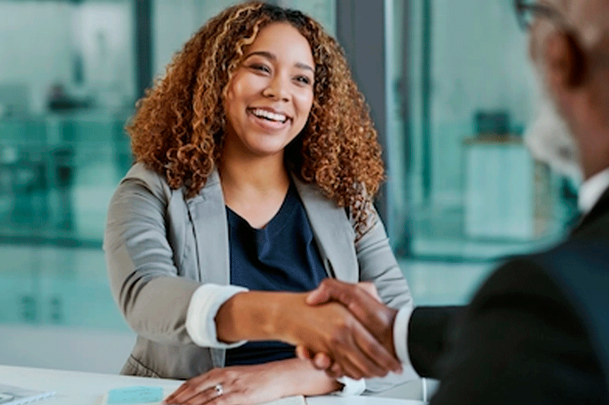 Mulher sorridente vestindo um blazer cinza aperta a mão de um entrevistador em um ambiente corporativo, transmitindo confiança e profissionalismo.
